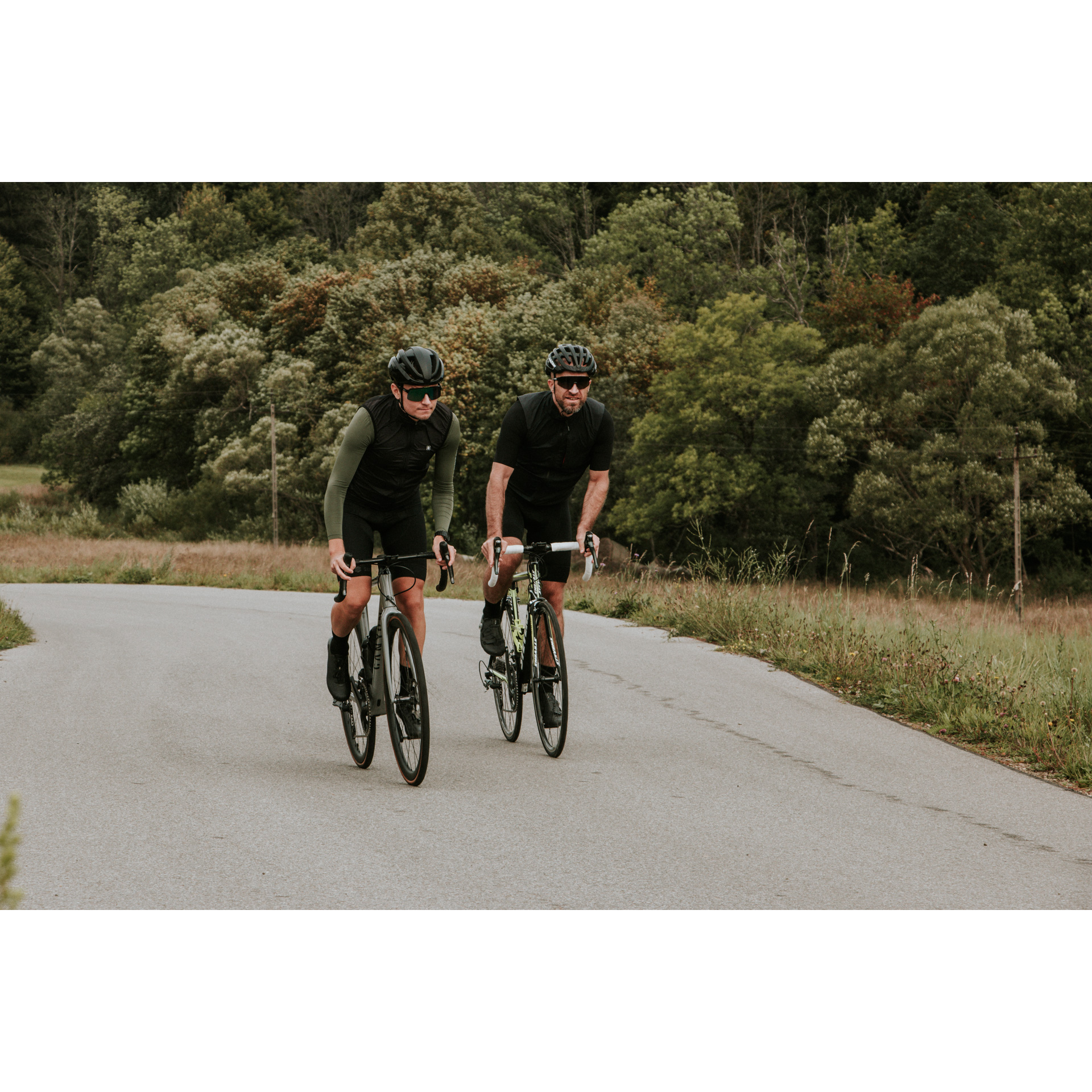 Dwójka rowerzystów w czarnych strojach i kaskach jadąca drogą asfaltową na tle zielonego lasu