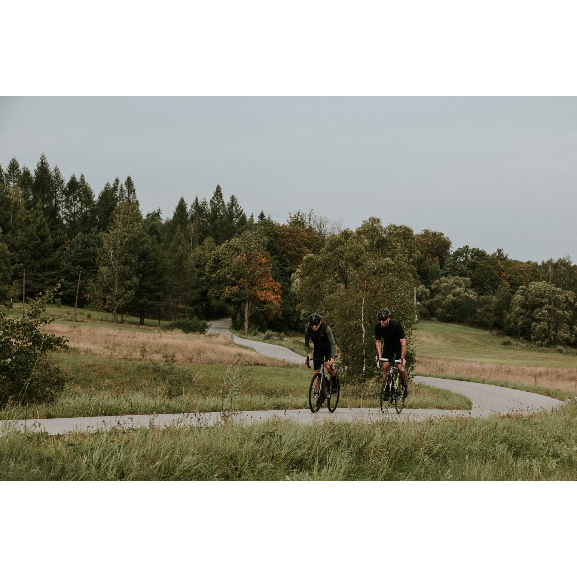 Dwójka rowerzystów w czarnych strojach wyjeżdżająca z zakrętu asfaltową drogą na tle zielonego lasu 