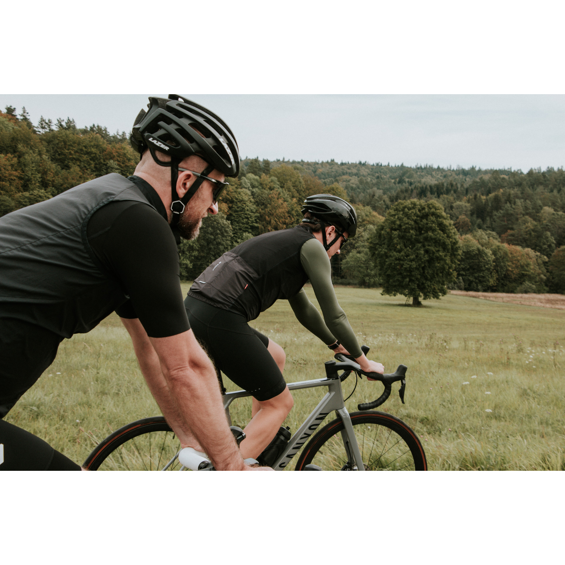 Dwójka rowerzystów w czarnych strojach i kaskach jadąca na rowerach na tle zielonej polany i gęstego lasu w oddali  