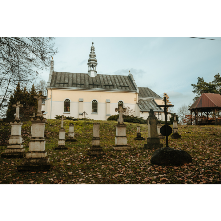 Krzyże i kamienne kapliczki cmentarne na tle jasnego kościoła z szarym dachem i wieżą