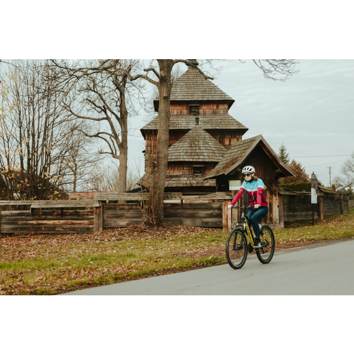 Rowerzystka w kasku i różowo-niebieskiej kurtce jadąca asfaltową drogą obok drewnianej cerkwi