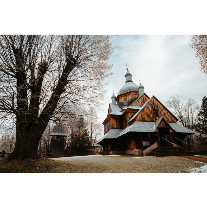 Drewniana cerkiew z jasnym dachem obok dużego, rozłożystego drzewa