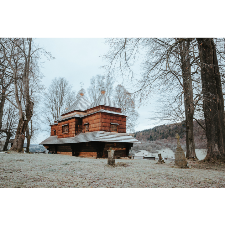 A wooden church with a snow-covered roof and a large window among trees and small stone crosses