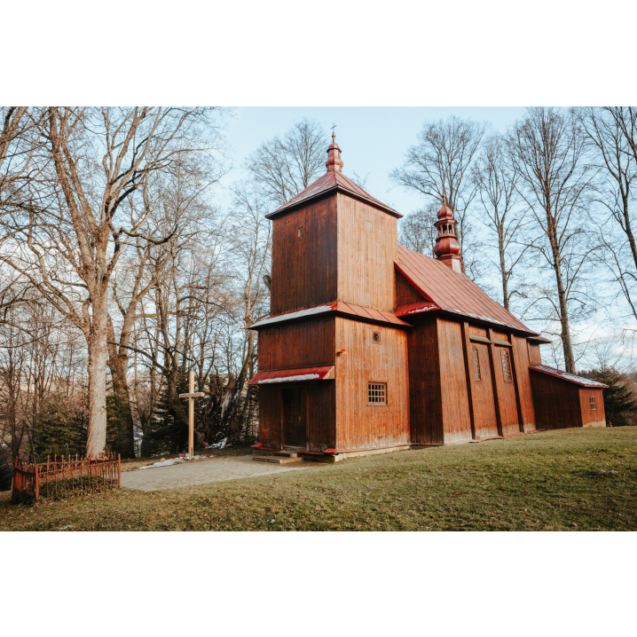 Drewniana cerkiew wśród wysokich drzew i dużego, drewnianego krzyża