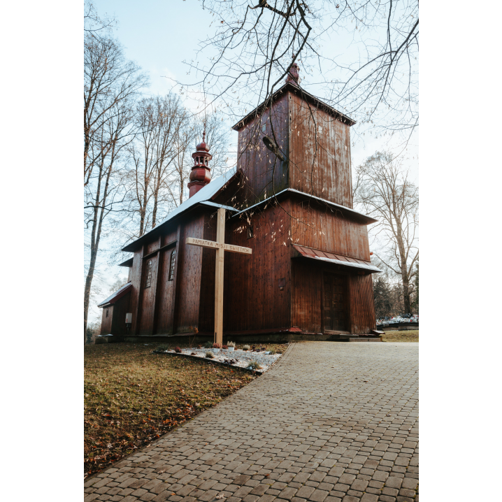 Ścieżka z kostki brukowej i wejście do drewnianej cerkwi wśród wysokich drzew i dużego, drewnianego krzyża