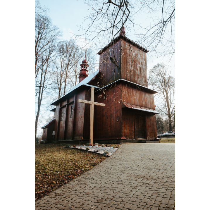 A path made of cobblestones and the entrance to a wooden church among tall trees and a large wooden cross