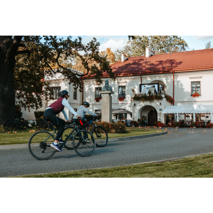 A woman and a child cycling along a paved road to a bright red-roofed manor house