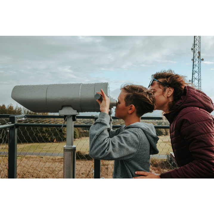 A boy in a gray sweatshirt and a woman in a maroon sweatshirt looking through large metal binoculars