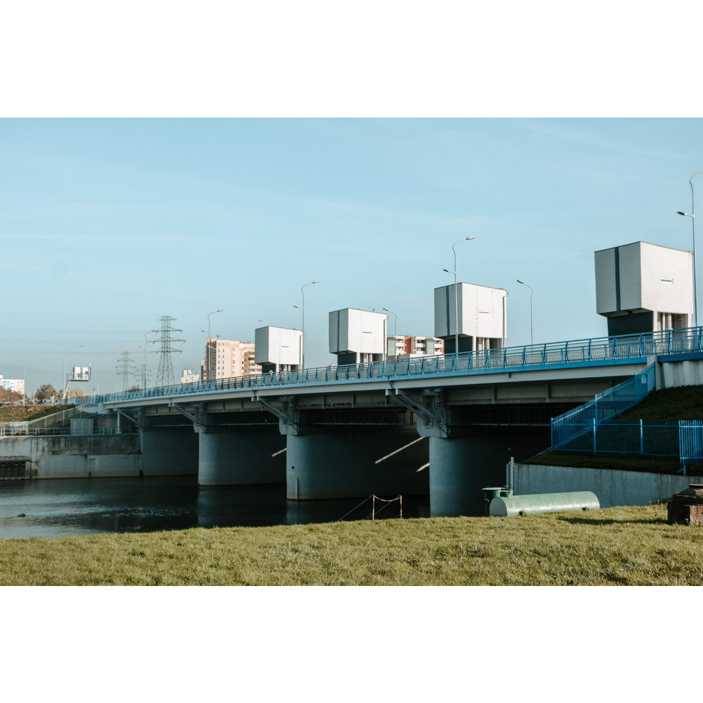 A river dam with three concrete pillars and four square towers visible, secured by blue barriers