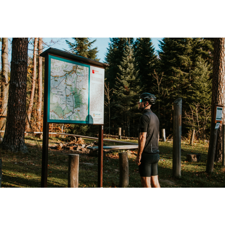 Rowerzysta w kasku ubrany na czarno patrzący na tablice informacyjną z mapą w lesie