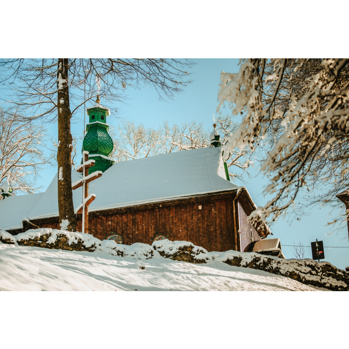 Drewniana cerkiew z zielonym dachem i wieżyczką z krzyżem obok drzewa i prawosławnego krzyża stojącego w śniegu