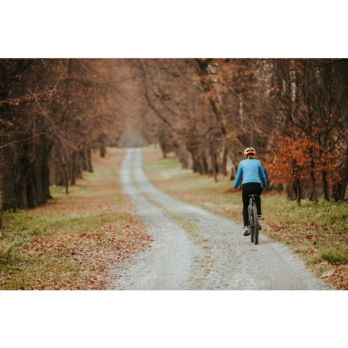 Rowerzystka w niebieskiej kurtce jadąca drogą wśród drzew z poboczem pokrytym jesiennymi liśćmi