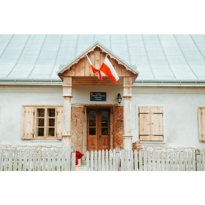 Wejście do murowanego niskiego domu z drewnianymi okiennicami, flagą Polski i szarym dachem otoczony białym, drewnianym płotem