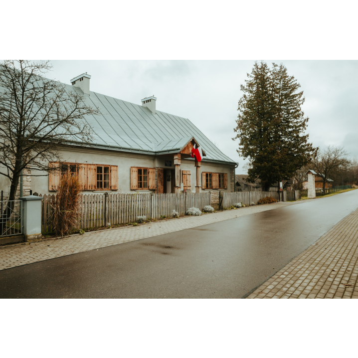 Murowany niski dom z drewnianymi okiennicami, flagą Polski i szarym dachem otoczony białym, drewnianym płotem przy asfaltowej drodze z chodnikiem