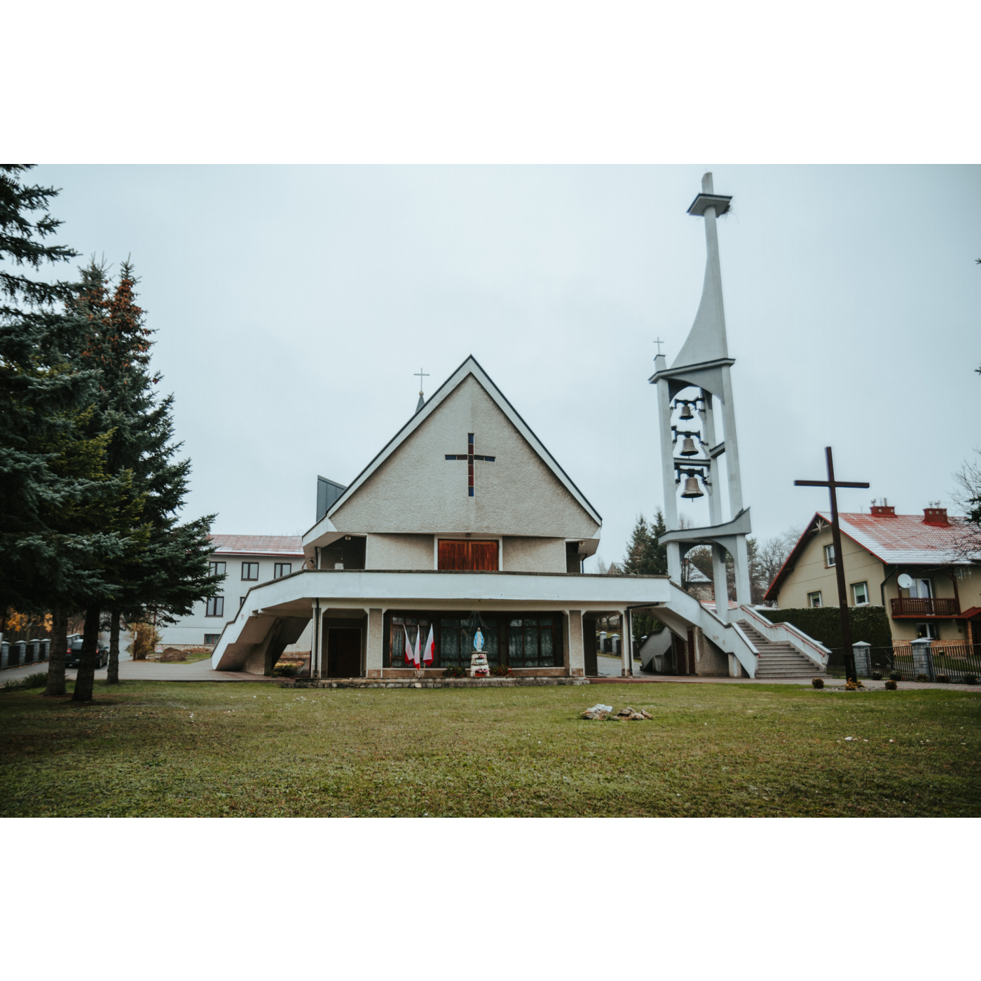 A church with a triangular façade and a cross and a statue of Our Lady under the stairs next to a tall belfry with three bells