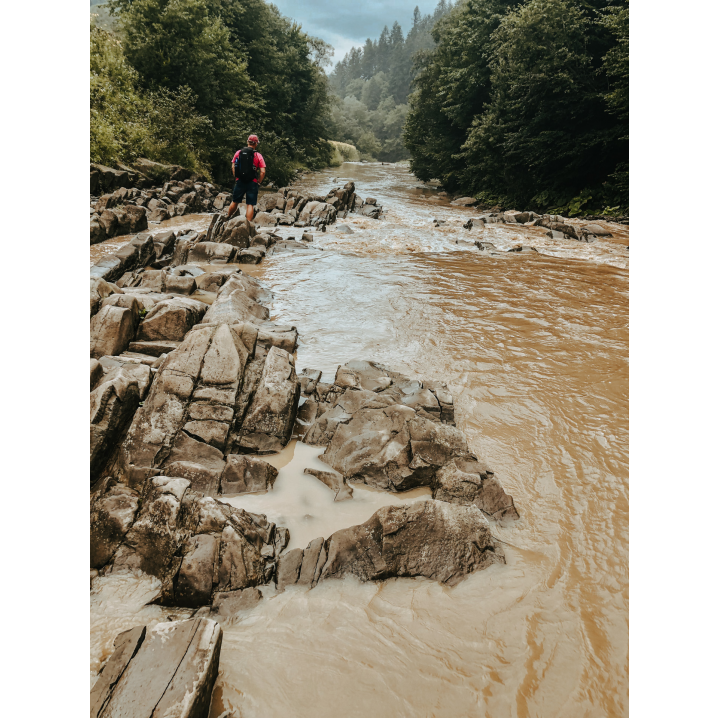 A man with a black backpack and a red cap standing on a stone in the river against the background of trees