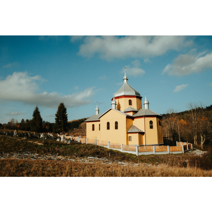 A wooden Orthodox church with a gray roof surrounded by a fence with a cemetery in the background