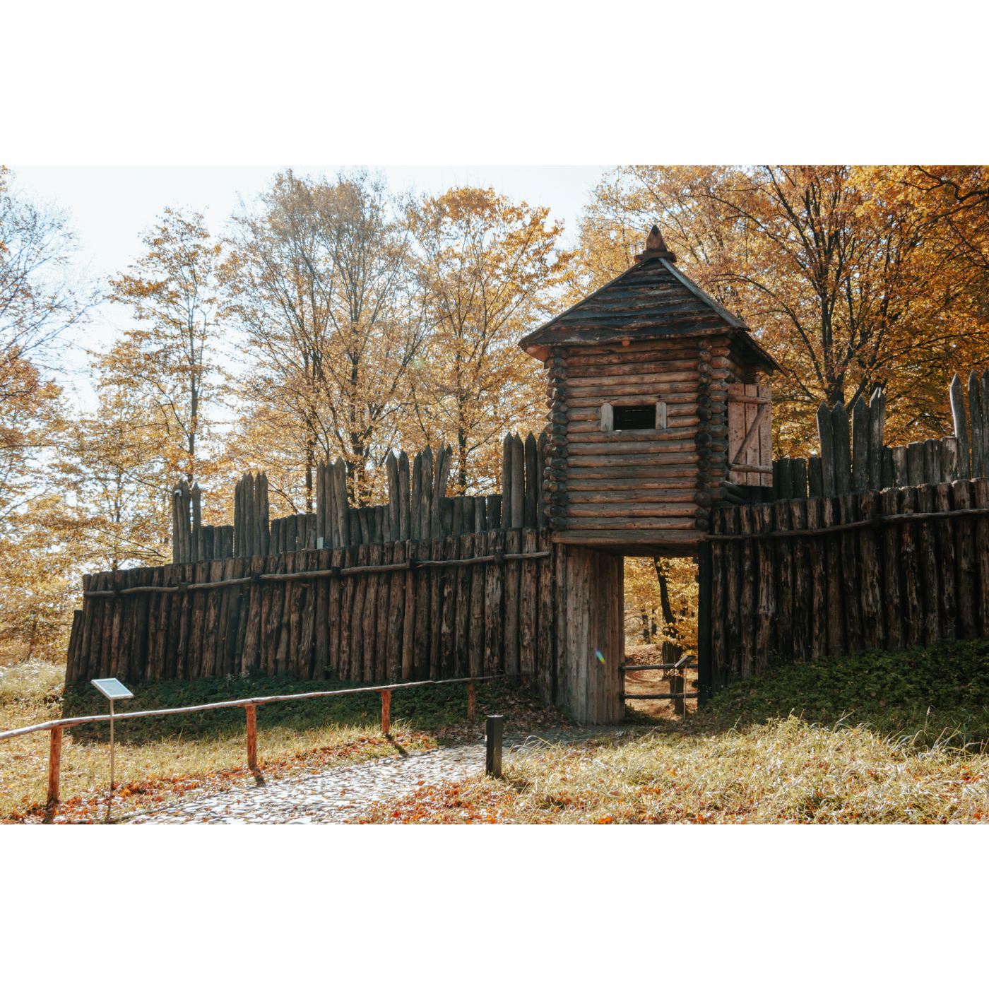 Entrance to a wooden castle with a fence made of wood piles against the background of autumn trees