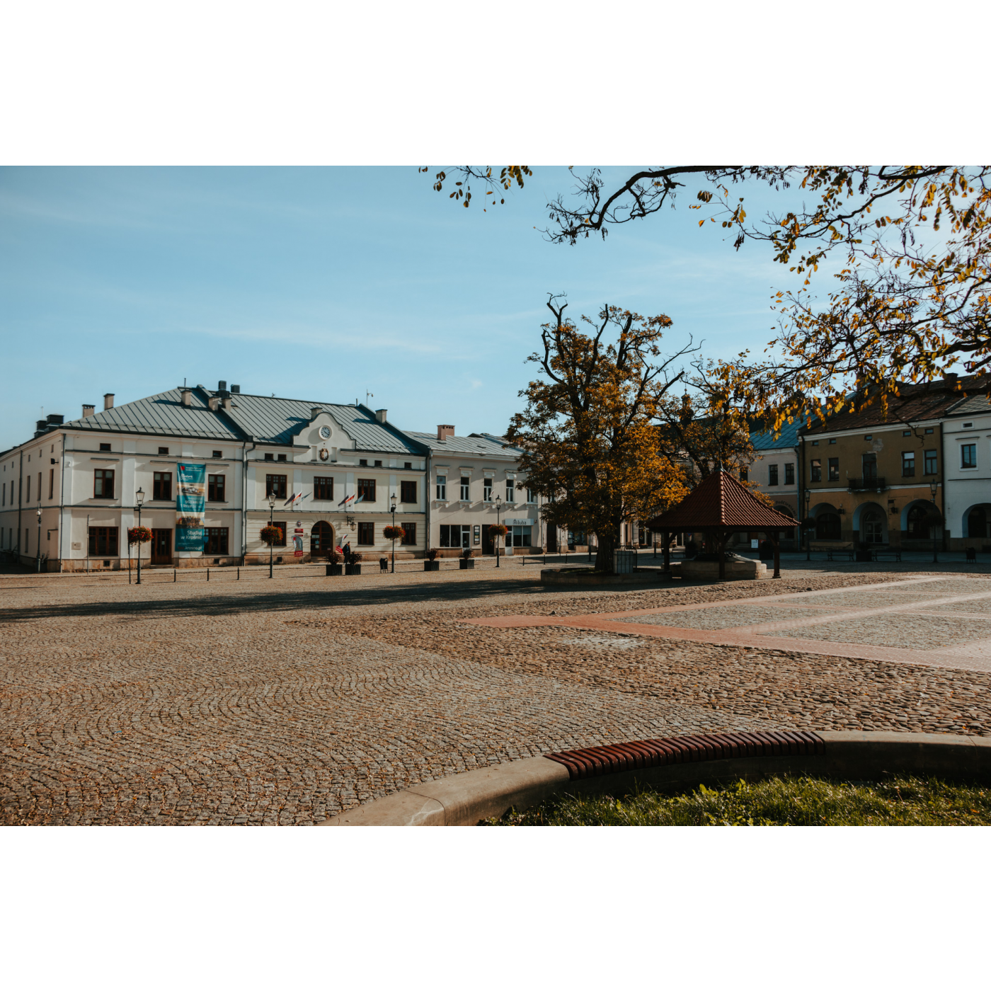 Market square with a view of a large white tenement house, urban buildings and a well standing by an autumn tree