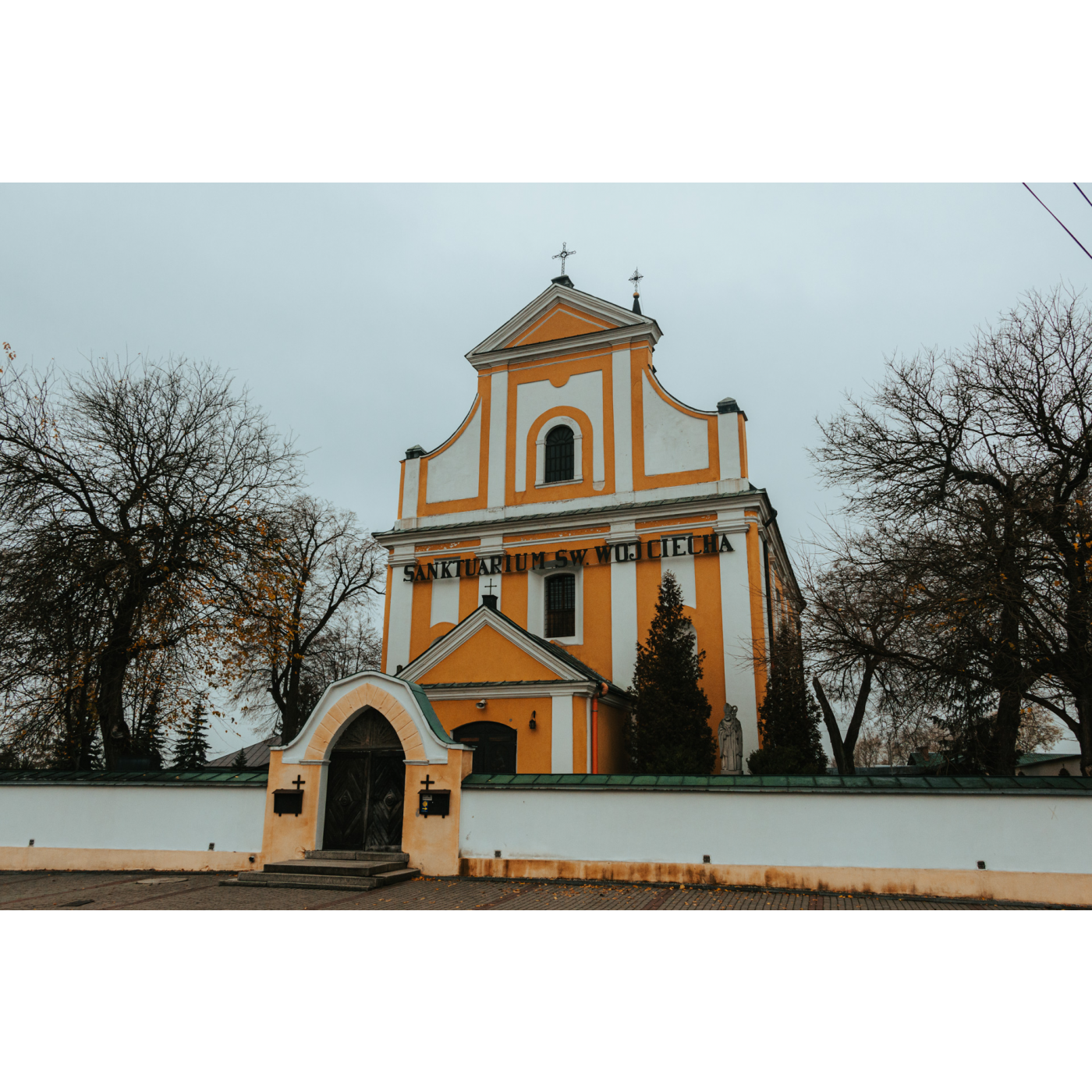 A white-orange church with an ornate facade and a black inscription fenced with a light-colored wall with an orange brick gate