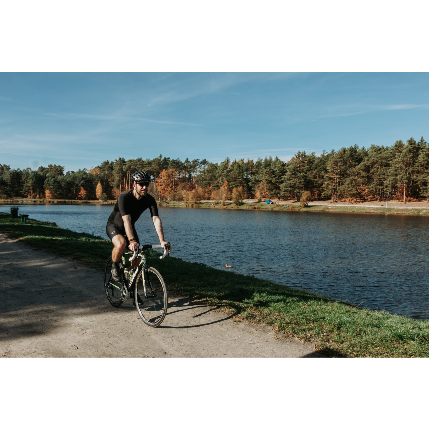 Rowerzysta w czarnym stroju i kasku jadący po drodze wzdłuż rzeki z zalesionym brzegiem