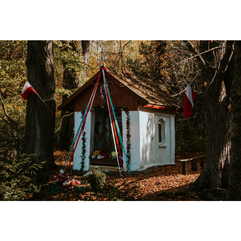 A white chapel in the forest, with a wooden roof, decorated with flowers and colorful ribbons and Polish flags