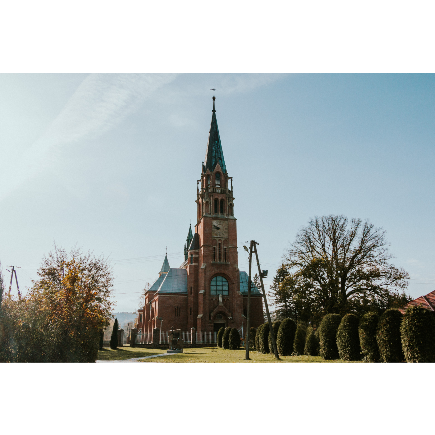 A brick church with a soaring tower and a clock on it, standing among trees and green bushes