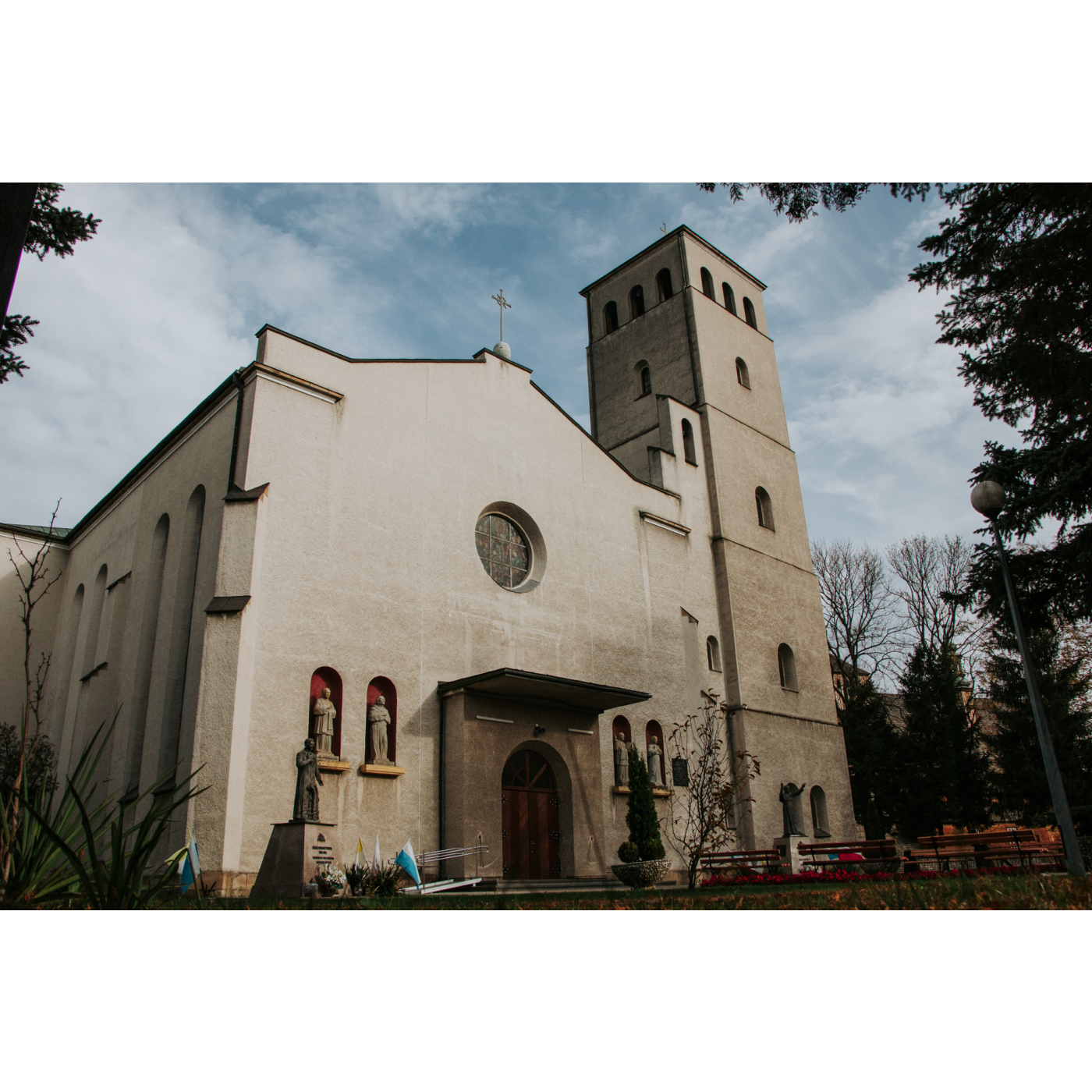 Murowany kościół w kolorze szarym o bardzo szerokiej fasadzie i wysokiej wieży, po obu stronach wejścia znajdują się figury świętych 