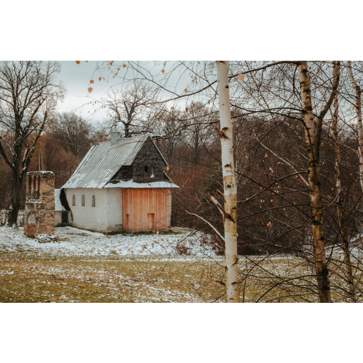 A small brick orthodox church with wooden elements on the front of the building, slightly covered with snow, shown from behind the trees surrounding the area