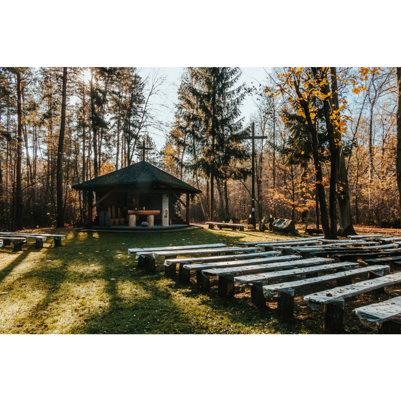 A small chapel made of wooden logs surrounded by autumn trees, wooden benches for the faithful in front, and a large wooden cross in the background