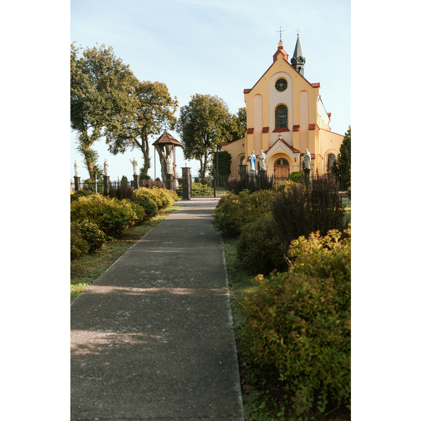 The yellow facade of the church with a soaring roof, a statue of the saint in front of the building, a tree on the left side