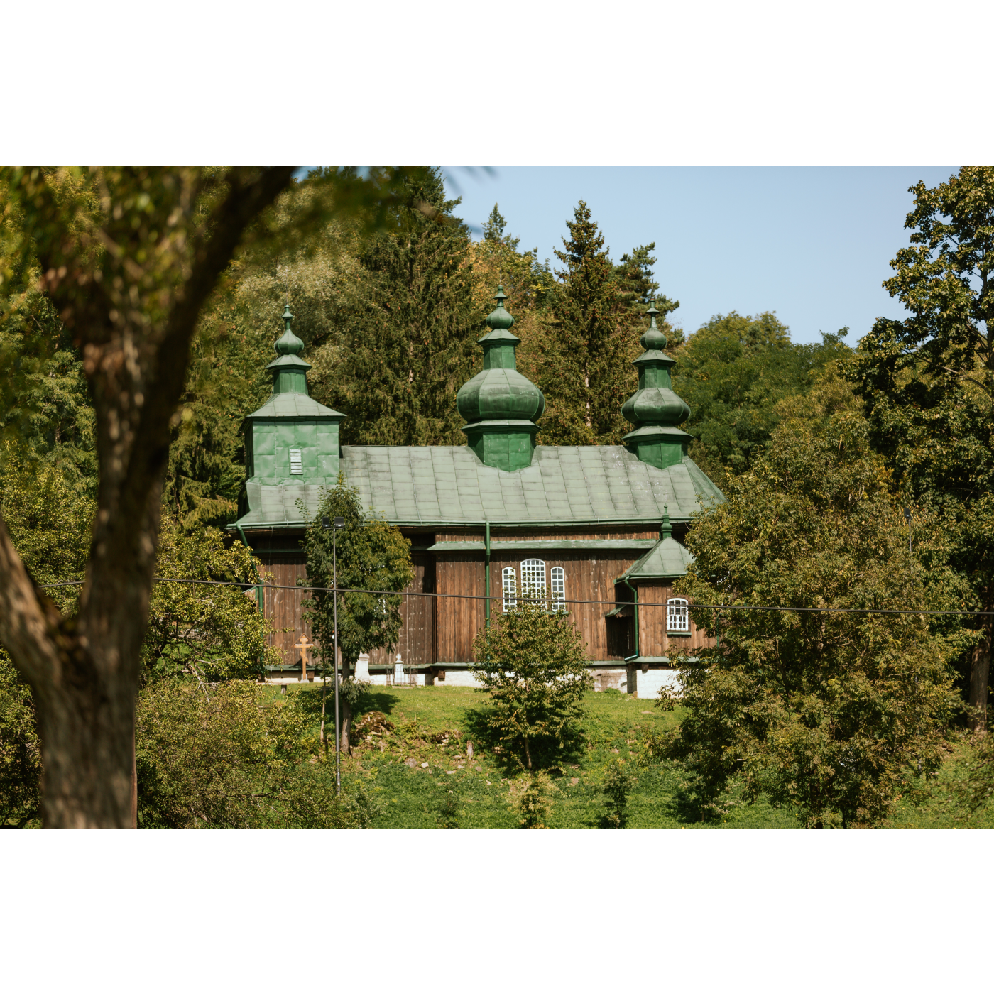 Drewniana cerkiew z zielonym dachem zwieńczonym trzema kopułami stojąca wśród zielonych, wysokich drzew