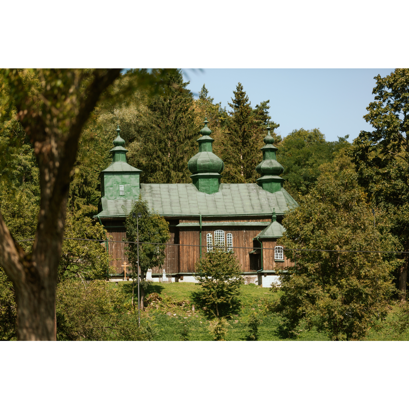 A wooden Orthodox church with a green roof topped with three domes standing among green, tall trees