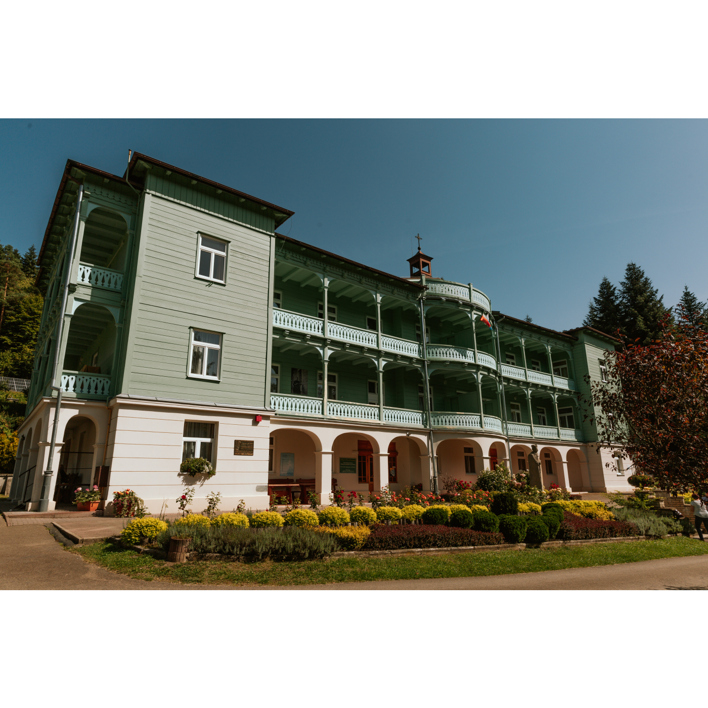 A three-story white and green building with a wide facade and shaded balconies on each floor.