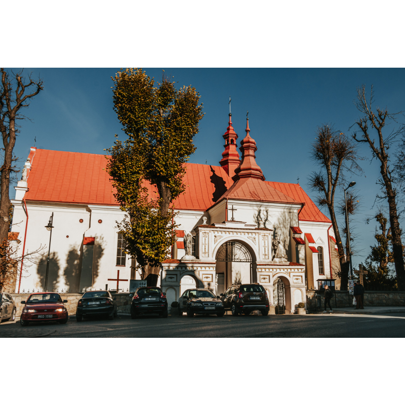 A bright brick church with a red roof and turrets and an entrance gate with ornaments and statues of saints