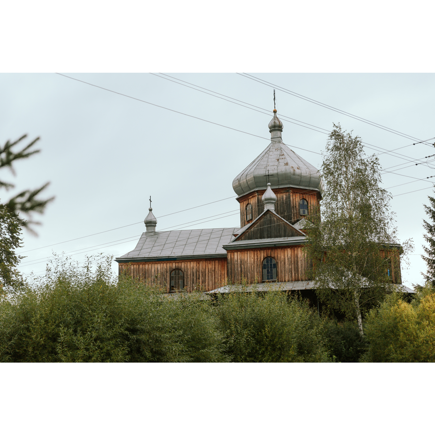 A wooden church with one main dome and a gray roof, green trees around